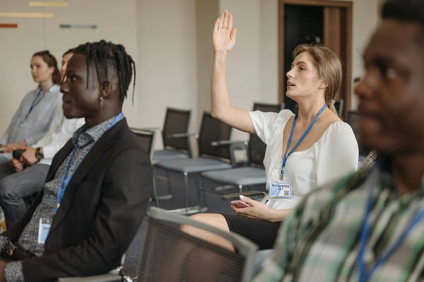 A woman raising her hand during a conference, with several other attendees seated around her.